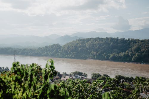 From above of picturesque scenery of green village and forest on hilly coasts of river against cloudy sky on sunny day