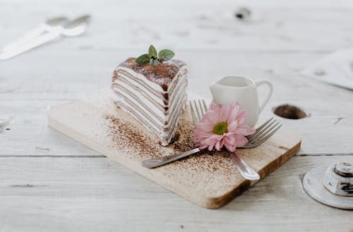 Free From above of appetizing piece of cake decorated chocolate powder and mint leaves served near ceramic creamer and forks with light pink chrysanthemum on top placed on wooden board Stock Photo