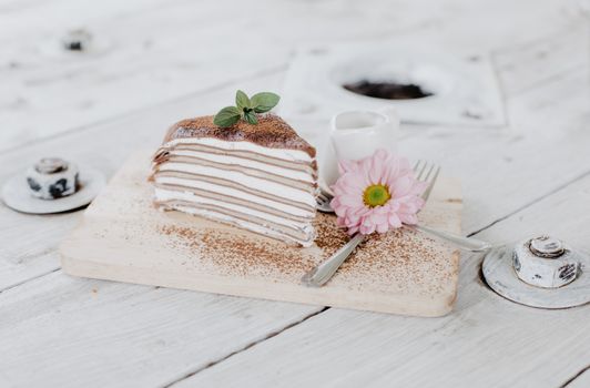 Slice of tasty layer pancake pie with sweet whipped cream placed on white wooden board near ceramic jigger and decorated with flower