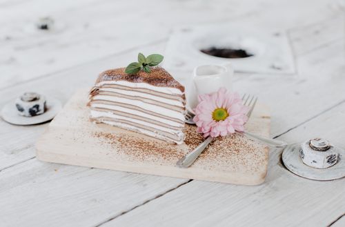 Free Slice of tasty layer pancake pie with sweet whipped cream placed on white wooden board near ceramic jigger and decorated with flower Stock Photo