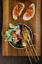 Top view of appetizing Asian seasoned meat skewered and grilled on sticks placed in pot with fresh sliced vegetables and served on wooden board with sandwiches topped with chopped tomatoes