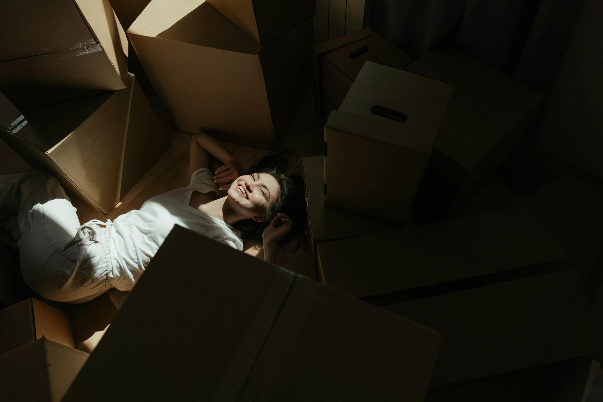 Smiling woman lying on floor surrounded by moving boxes, bathed in natural light.
