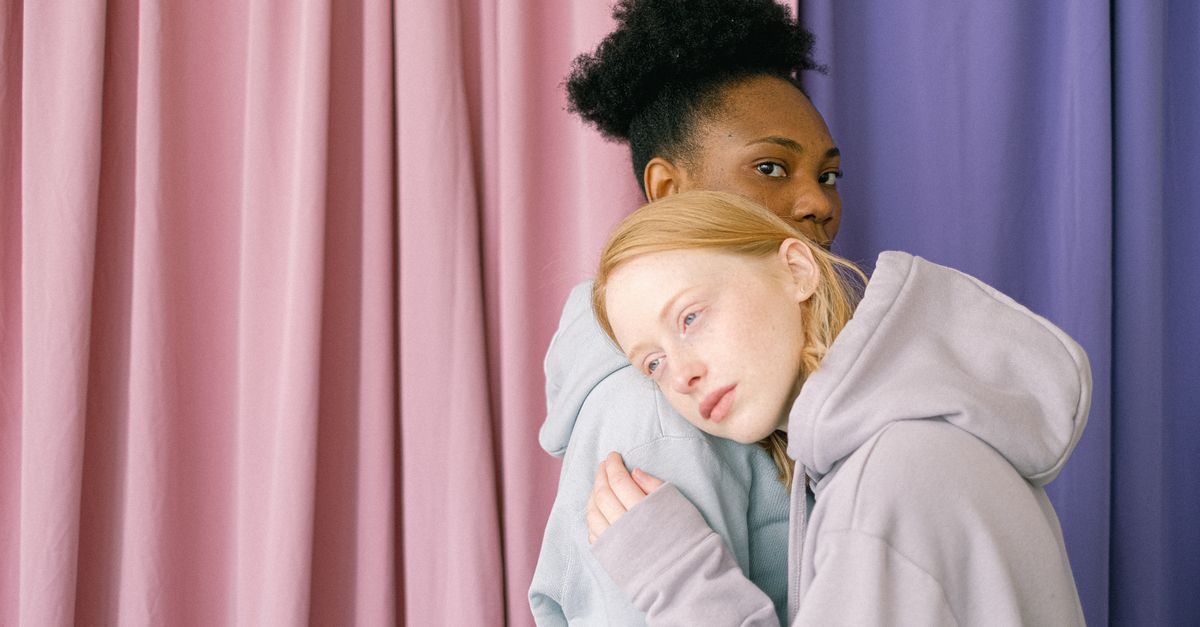 Interracial lesbian couple embracing indoors against pink and purple curtains.