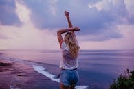 匿名女性穿着牛仔短裤和白色上衣站在沙滩上，举起双臂，欣赏傍晚海景和紫色天空的风景如画