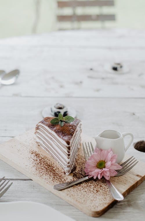 From above composition of sweet pancake cake with whipped cream placed on light wooden board with cutleries and delicate pink flower