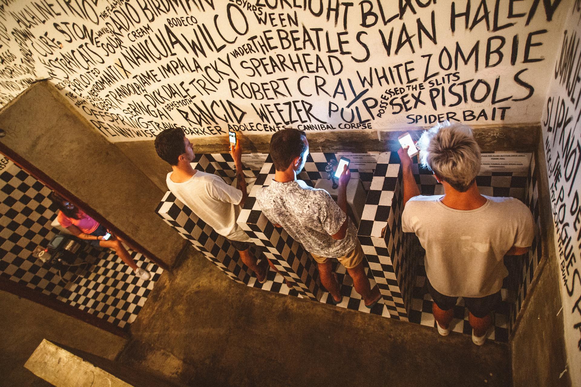 From above full length faceless people in casual clothes using mobile phones in public toilet with black and white tiles and inscriptions on walls gadget addiction concept