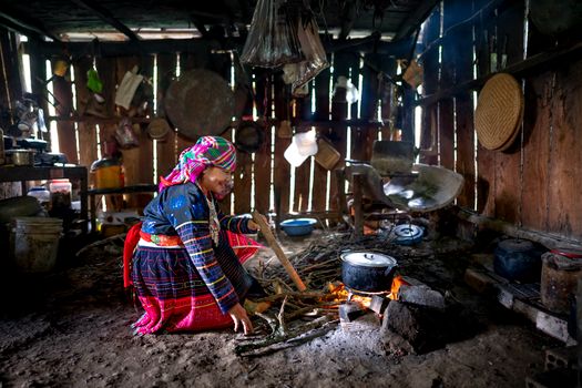 Full body side view of smiling adult ethnic female in colorful national dress and headscarf cooking soup on open fire while sitting on ground in simple wooden construction