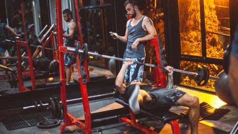 Full body concentrated sporty fitness trainer using smartphone while male on bench struggling with heavy barbell in fitness club gadget addiction concept