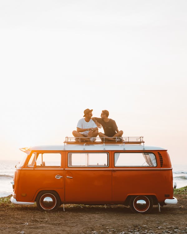 Silhouettes of full body faceless male friends in casual clothes resting on camper van roof and chatting in countryside in sunset time