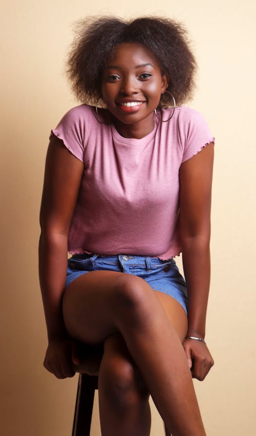 Free African American woman in casual shorts and t shirt with short dark curly hair sitting on chair and looking at camera with toothy smile Stock Photo