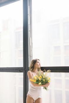 Caucasian woman in lingerie holding a bouquet of yellow tulips by a large glass window.