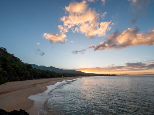 Breathtaking scenery of tranquil shoreline with green trees and mountains on background under cloudy sky at sunset