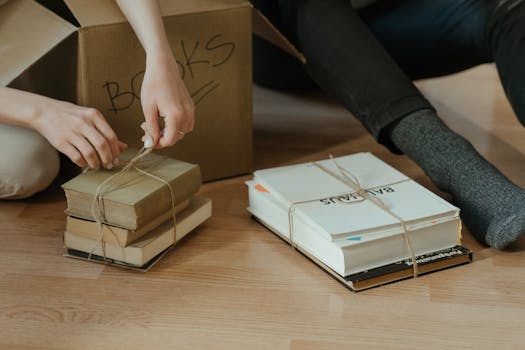 Hands organizing books on the floor next to cardboard box. Ideal for themes of moving and home organization.