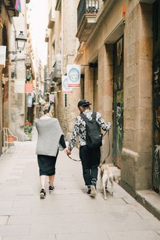 一对夫妇手牵着手，在一条狭窄的、两旁环绕着历史建筑的街道上遛狗
