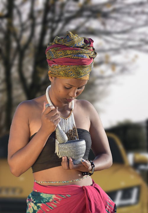 Young lady in traditional outfit stir something in ceramic bowl while standing on street against modern yellow car