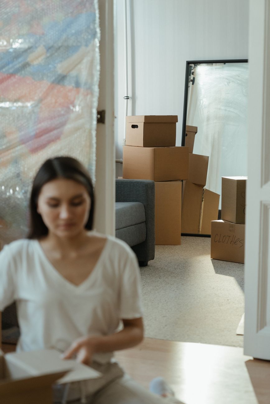 A woman sitting among unpacked boxes in a bright apartment, suggesting the unstable middle ground between leaving and settling.
