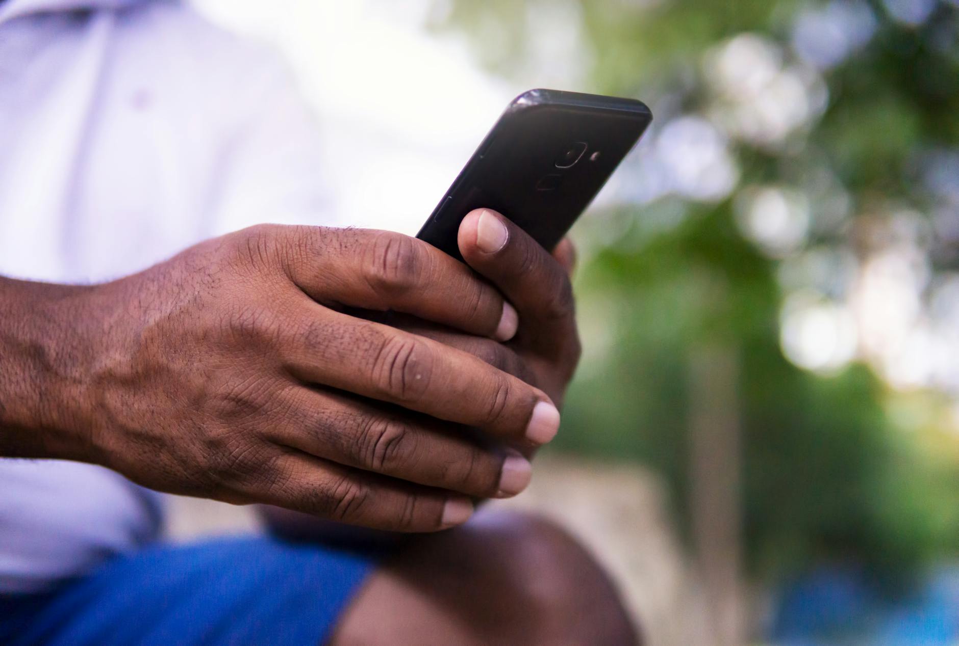A close-up of a man's hands holding a smartphone in a park setting
