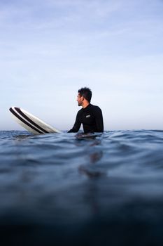 一名穿着黑色潜水服的短发男性冲浪者，正低角度坐在冲浪板上，目光移开，望向平静的蓝色大海