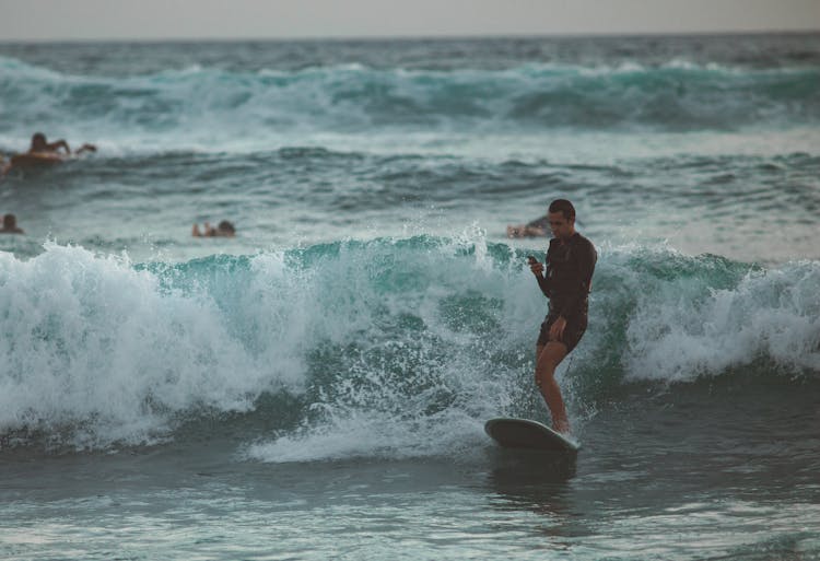 Young Male Surf Rider Surfing On Board And Using Smartphone