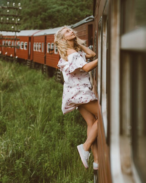 Free Side view of full body young woman in air dress leaning out from exit door and holding on to railroad while riding train past green grass Stock Photo