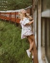 Side view of full body young woman in air dress leaning out from exit door and holding on to railroad while riding train past green grass