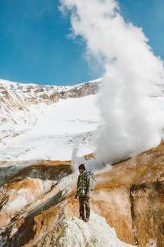 一名身穿保暖外套、背着背包的男性旅行者站在粗糙的山顶，靠近火山喷出的蒸汽，紧贴蓝天下的积雪山坡
