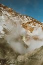 Rough mountain slope covered with stones and snow with volcanic steams blowing off in air under blue sky