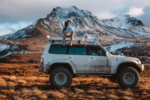 Full body woman in casual clothes and hat sitting on offroader rooftop against snowy mountain range in sunny cloudy day