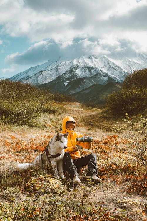 Full body frozen traveler resting on grassy highland with adorable Husky dog and pouring hot drink from thermos bottle against majestic snowy mountain range on cold day