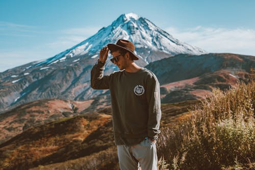 Young stylish man in hat and modern sunglasses standing on hill with hand in pocket against high snowy mountain