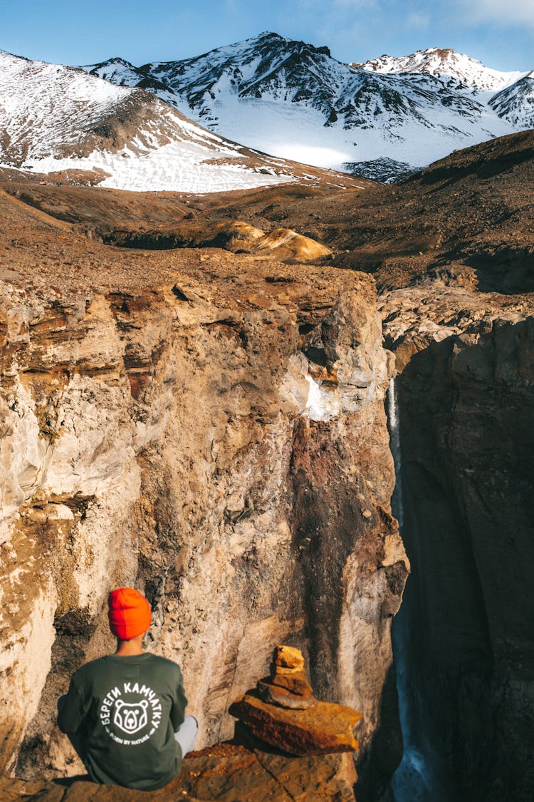 Unrecognizable Hiker Sitting On Rock Above Gorge Against Snowy Mountain Slopes