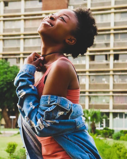 Side view of black woman in denim jacket smiling while standing in city near multi storey building during summer day