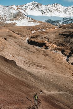在晴朗的日子里，徒步旅行者面目全非，行走在狂野的河流与雪山附近的火山地形之间的狭窄小路上