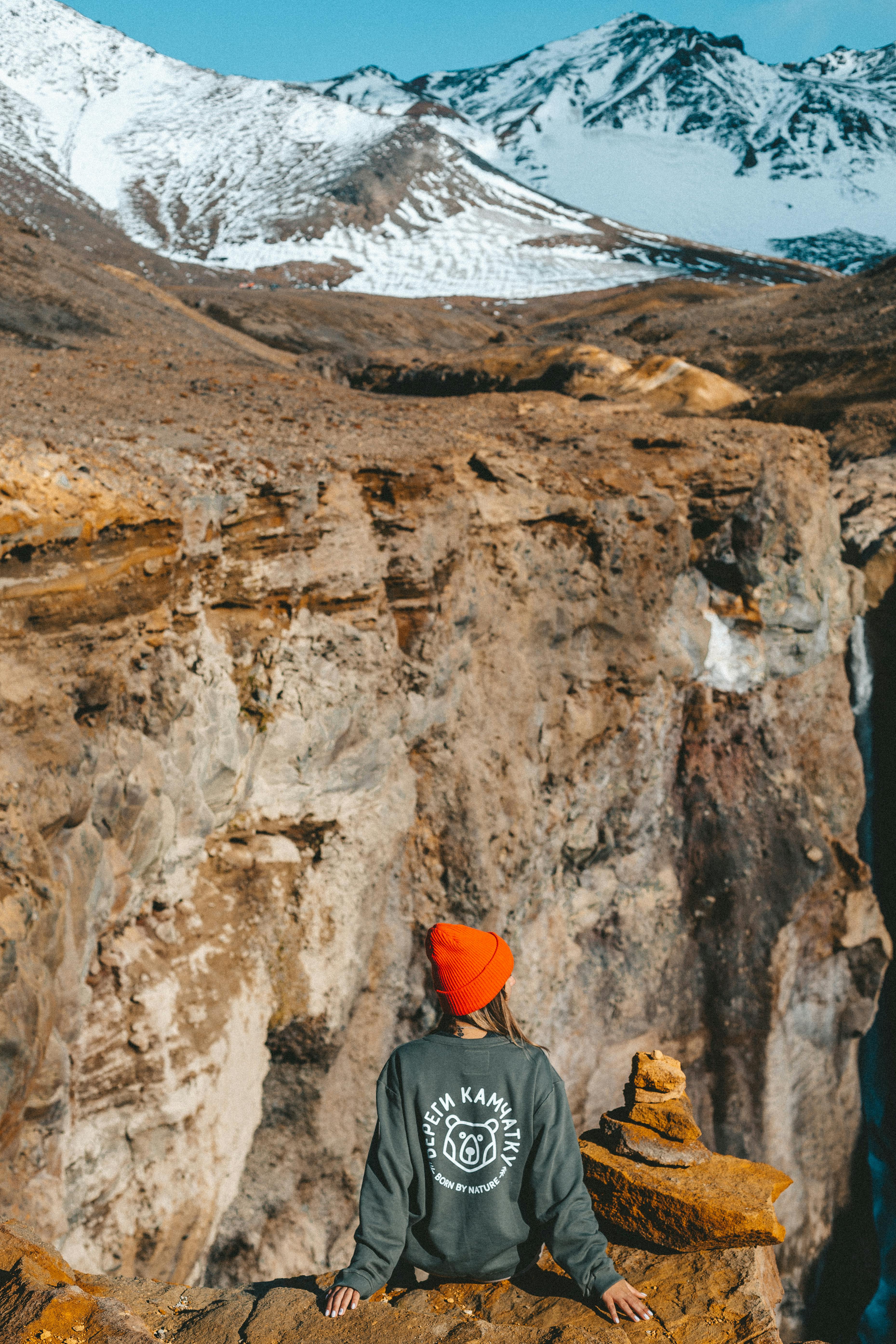 Back view of anonymous female in knitted hat and warm sweatshirt admiring stunning beauty of snowy mountains and rocky terrain while sitting on cliff edge of Dangerous Canyon in Kamchatka