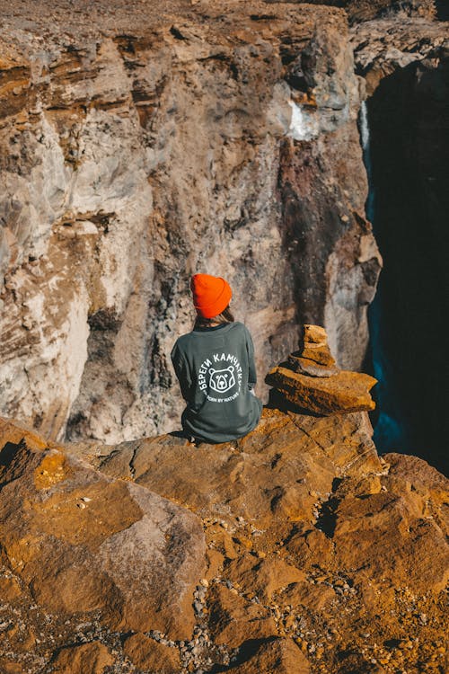 Back view of lonely female in orange hat sitting on edge of canyon and admiring picturesque view