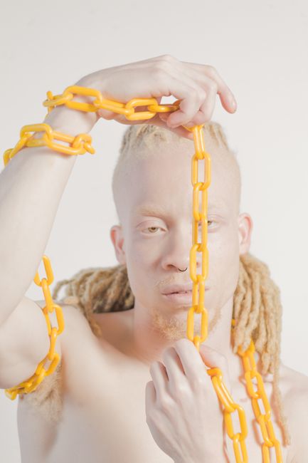 Albino male model with dreadlocks holding and posing with yellow chains, indoors.