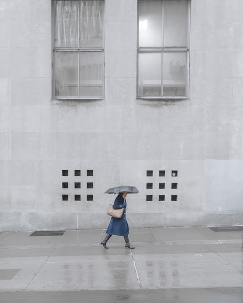 Full body of anonymous female in distance strolling with umbrella on pavement near building with gray facade during rainy weather