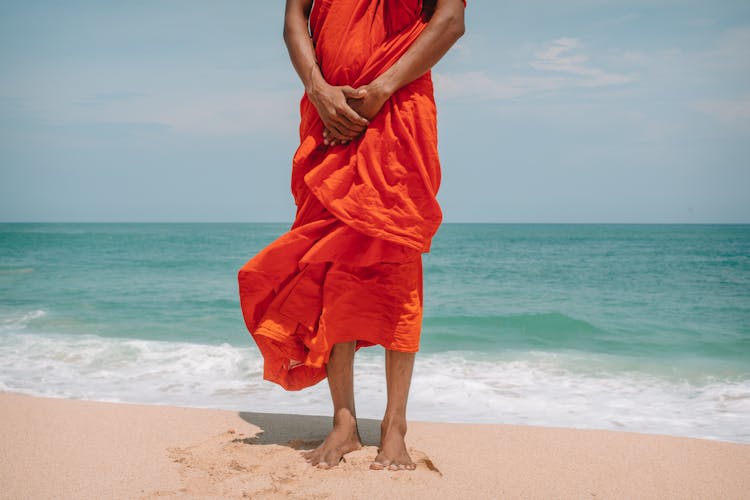 Crop Ethnic Man In Orange Robe Standing On Sandy Beach