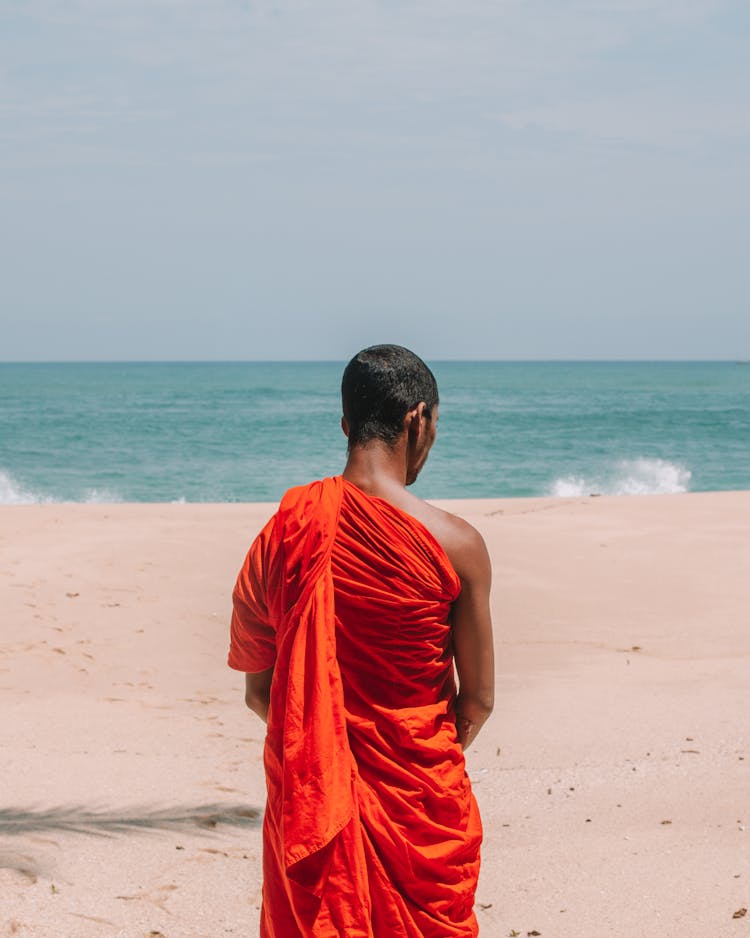 Unrecognizable Asian Man In Traditional Orange Robe On Beach