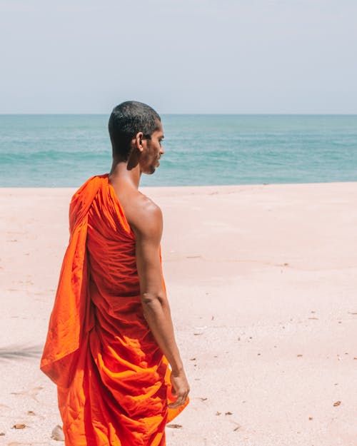 Side view of young Asian male Buddhist monk in traditional orange dress walking on sandy shore and admiring calm sea under clear blue sky