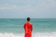 Unrecognizable Asian male monk in traditional orange robe on seashore