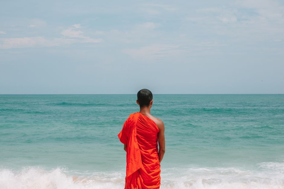 Back view of young barefoot ethnic male Buddhist monk standing on sandy beach near waving ocean during serene sunny day