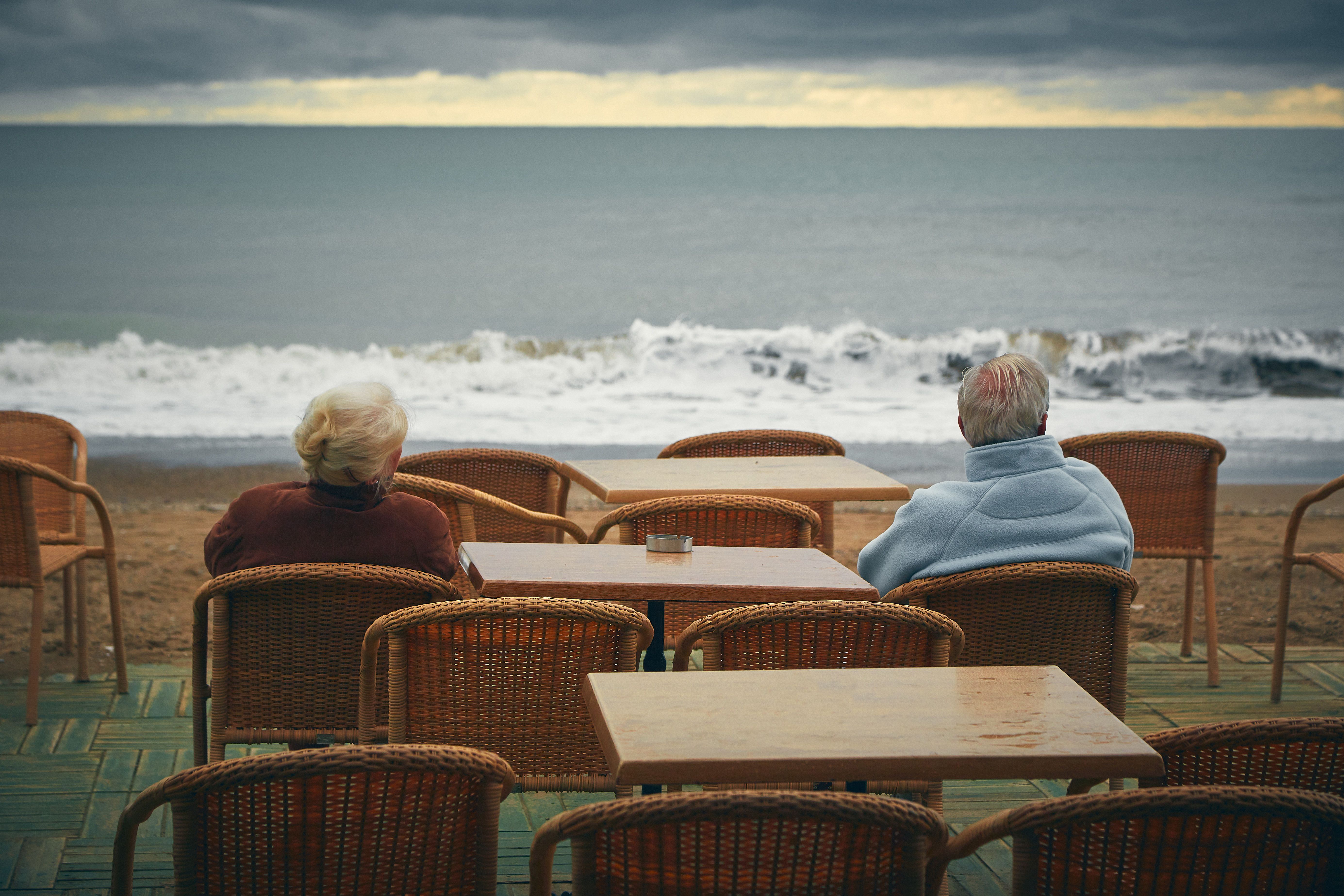 Back view of anonymous elderly man and woman contemplating wavy sea with horizon line while sitting at table in cafeteria near beach in evening