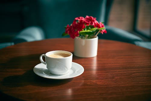 From above of ceramic cup of coffee on saucer near small vase with bright artificial flowers on wooden table at home