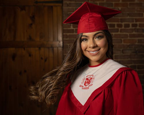 Positive young female in graduation clothes standing near brick wall and looking at camera with toothy smile