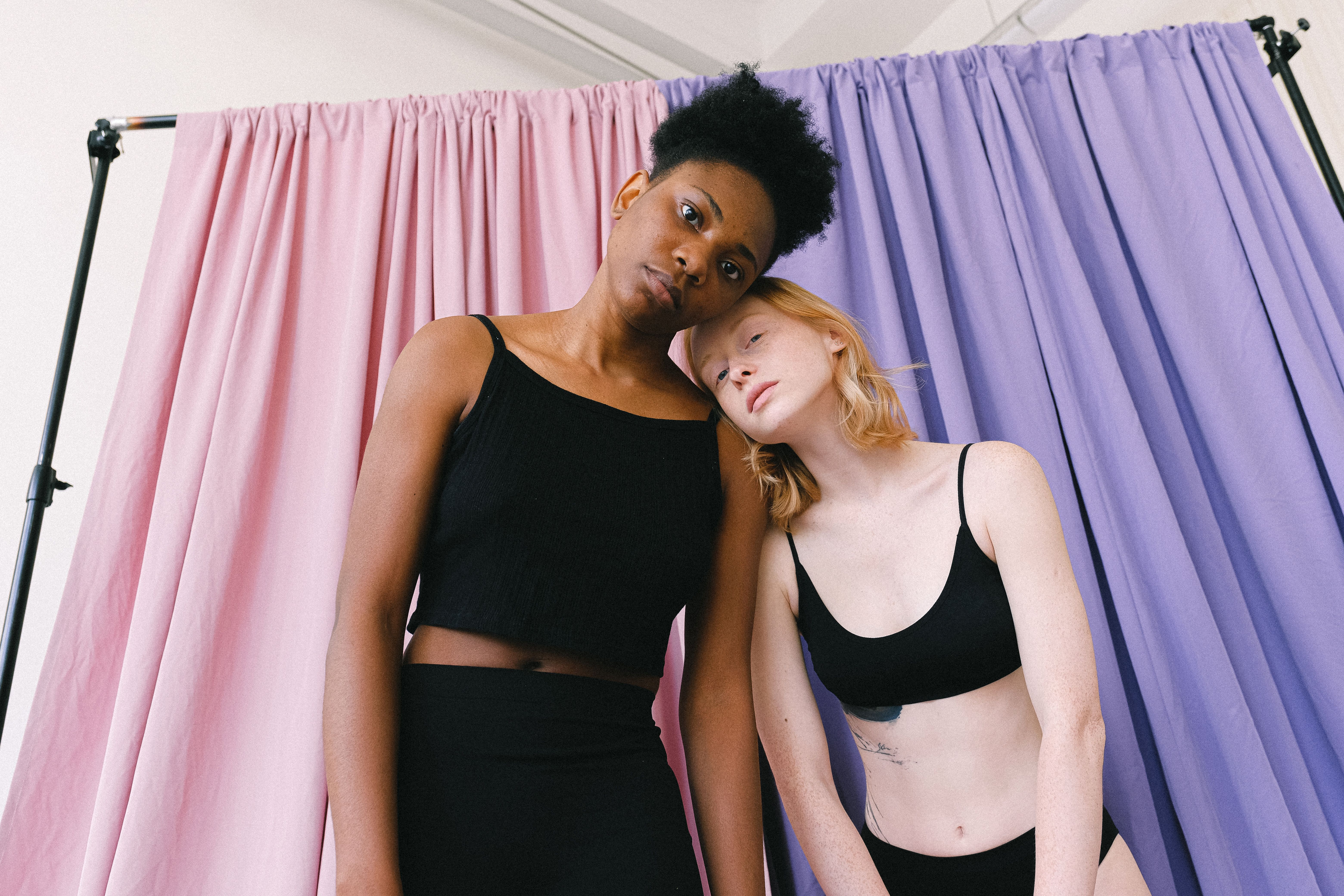 Two women in sportswear pose with affection against pink and purple curtains, showcasing diversity.