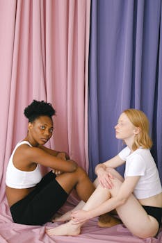 Two women in casual wear sitting barefoot on the floor with pink and purple curtains in the background.