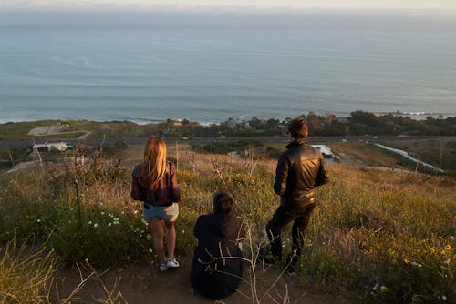 From above back view of young friends in casual outfits observing picturesque seascape from high edge of hill in rural area while enjoying weekend together