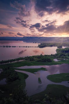 A peaceful sunset over a serene river and wetland landscape with dramatic clouds overhead.