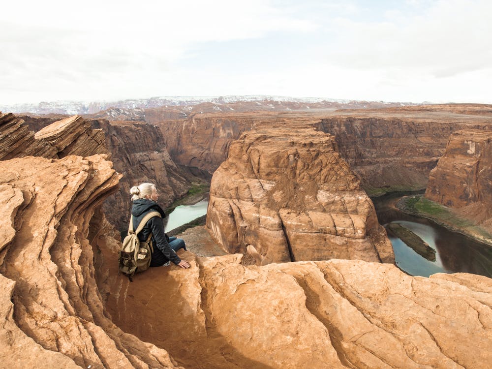 Free Back view of young female tourist in warm jacket with backpack sitting on edge of rock and observing spectacular scenery of canyon with river against cloudy sky Stock Photo